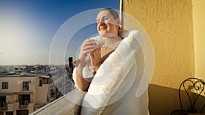 Beautiful smiling woman drinking tea on the terrace and looking at sunrise over the city. People in morning, relaxing at home,