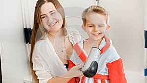 Beautiful smiling woman drying hair and blowing hot air with hairdryer on her little son after washing in bath. Concept