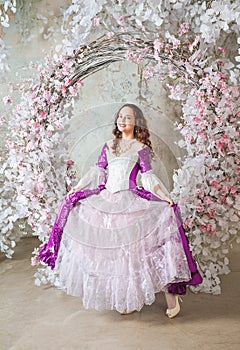 Beautiful smiling woman in fantasy white and purple rococo style medieval dress standing near decoration with white flowers and