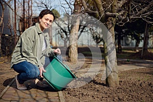 Beautiful smiling woman gardener waters tree from a bucket, enjoying gardening on beautiful sunny spring day. Springtime,