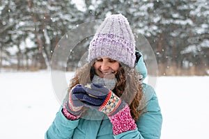 Beautiful smiling woman with long curly hair in blue winter jacket and knitted colorful gloves is holding in her hands cup of hot