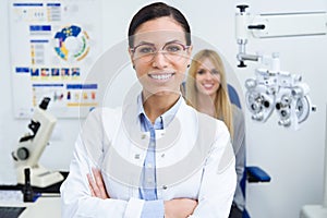 Beautiful smiling woman looking at camera in ophthalmology clinic with woman patient sitting for eye test in background.