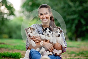 A beautiful smiling woman with a ponytail and wearing a striped shirt is holding three sweet husky puppies on the lawn