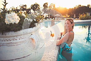 Beautiful smiling woman in the pool with cocktail. Warm lights and intense evening sun