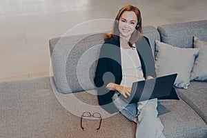 Beautiful smiling woman with red hair sitting with laptop and wireless headset on sofa in office