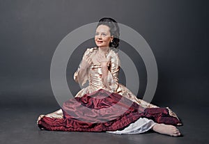 Beautiful smiling woman in rococo style medieval dress sitting on the floor against dark background