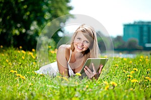 Beautiful smiling woman with tablet pc, outdoors. Beautiful young girl lying on the grass in the field. Smiling trendy stylish