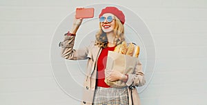 Beautiful smiling woman taking selfie picture by phone holding grocery shopping paper bag with long white bread baguette