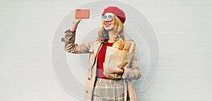 Beautiful smiling woman taking selfie picture by phone holding grocery shopping paper bag with long white bread wearing red beret