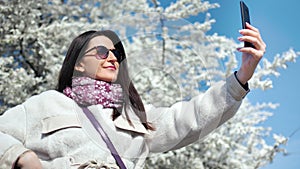 Beautiful smiling woman taking selfie using smartphone posing at white flowering sakura tree background