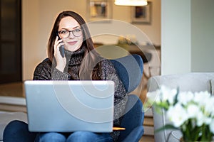 Beautiful smiling woman talking with somebody on mobile phone and using her notebook while sitting on chair at home
