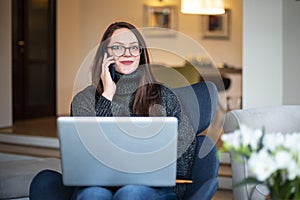 Beautiful smiling woman talking with somebody on mobile phone and using her notebook while sitting on chair at home