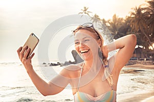 Beautiful smiling woman traveler in bikini on beach making selfie mobile photo on smart phone during beach holidays at beach for s