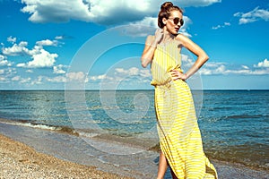 Beautiful smiling woman with updo hair wearing long yellow striped baggy summer dress and round sunglasses walking along the sea