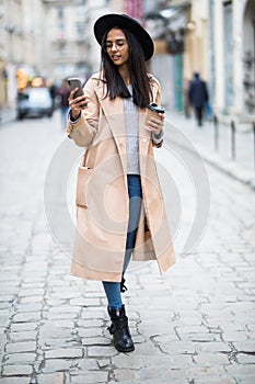 Beautiful smiling woman walking on city street from work with coffee cup and texting on mobile phone