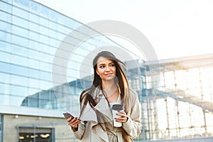 Beautiful smiling woman walking on near office from work with coffee cup and texting on mobile phone