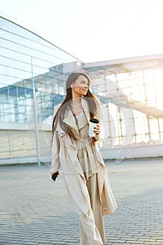 Beautiful smiling woman walking on near office from work with coffee cup and texting on mobile phone.Phone communication