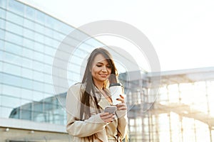 Beautiful smiling woman walking on near office from work with coffee cup and texting on mobile phone.Phone communication.