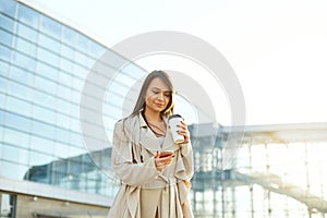 Beautiful smiling woman walking on near office from work with coffee cup and texting on mobile phone.Phone communication.