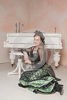 Beautiful smiling woman wearing green medieval vintage Victorian Style dress sitting on the floor near piano