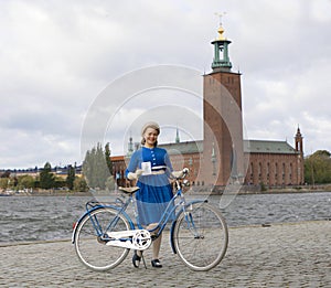 Beautiful smiling woman wearing old fashioned blue dress holding a retro bicycle in front of Stockholm City Hall