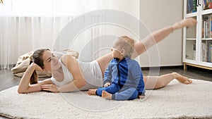 Beautiful smiling woman lying on carpet with baby son and doing fitness exercises. Family healthcare, active lifestyle, parenting
