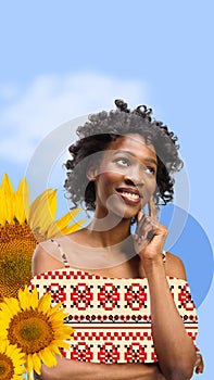 Beautiful, smiling, young african girl wearing clothes with Ukrainian ornament over blue background with sunflowers