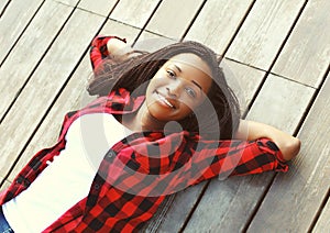 Beautiful smiling young african woman relaxed on wooden floor with hands behind head, wearing a red checkered shirt