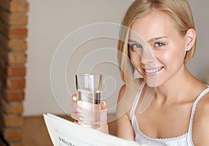 Beautiful smiling young blonde woman drinking water and reading newspaper in the morning