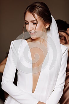 beautiful smiling young bride in long white satin dress with deep neckline, hairstyle and make up standing in hotel room
