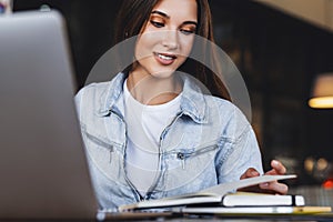 Beautiful smiling young business woman sits in front of laptop, makes notes in notebook.