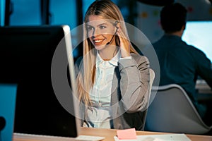 Beautiful smiling young business woman working with computer while talking with earphone sitting in the office