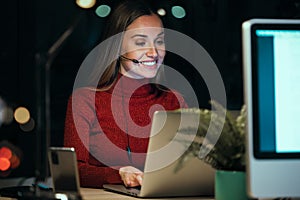 Beautiful smiling young business woman working with computer while talking with earphone sitting in the office at night