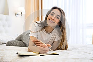 Beautiful smiling young girl lying on the bed writing or reading her book  in the morning