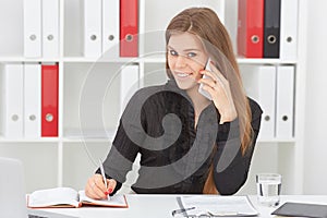 Beautiful smiling young girl make notes talking on the mobile phone sitting at office.