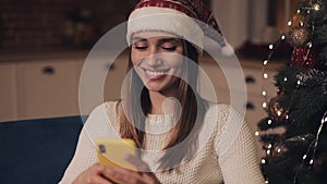 Beautiful smiling young lady wearing santas hat sitting on the sofa near christmas tree using her smartphone concept of