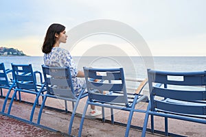 Beautiful smiling young mixed race woman sitting at one of the famous blue chairs at the Promenade des Anglais in Nice