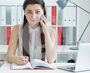 Beautiful smiling young secretary talking on the mobile phone sitting at office an looking into the camera. Business