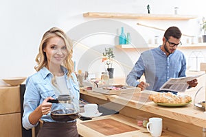 beautiful smiling young waitress holding coffee pot and cup while client reading