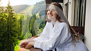 Beautiful smiling young woman in bathrobe looking on mountains and forest from the hotel room balcony