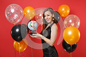 Beautiful smiling young woman in little black dress celebrating, holding world globe on bright red background air