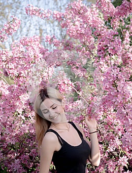 Beautiful smiling young woman near the blossoming spring tree. Portrait of pretty blond girl with long hair in pink flowers.