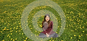 Beautiful smiling young woman relaxing on a meadow with many dandelions in the spring sun. Frontal view with copyspace.