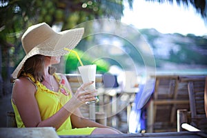 Beautiful smiling young woman in sunglasses wearing hat with a cocktail at the beach cafe