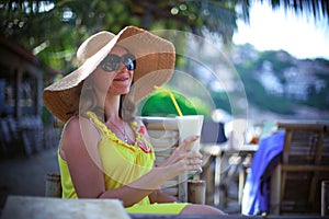 Beautiful smiling young woman in sunglasses wearing hat with a cocktail at the beach cafe