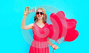 Beautiful smiling young woman taking selfie picture by phone with pink heart shaped air balloons on colorful blue