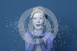 Beautiful Snow Queen woman with white creative makeup blowing snowflake on blue background. Carnival or Halloween party makeup.