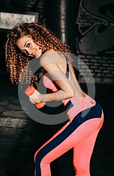 Beautiful sportive woman with blond curly hair in elegant sportive suit trains in gym with dumbbells