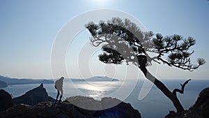 Beautiful sports girl with a backpack climbs on a high rock and looks at a beautiful view of the sea with the sun