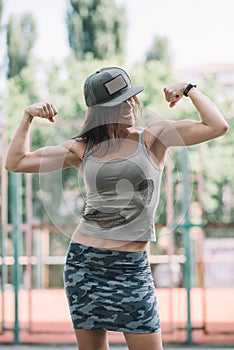 Beautiful sports girl in khaki clothes and a cap on the playground shows biceps and smiles. Healthy lifestyle.